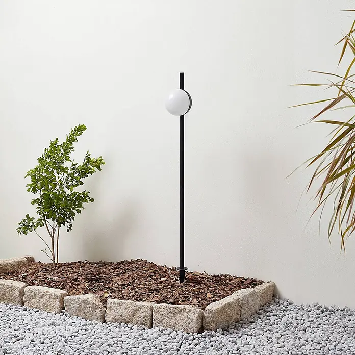 Lindby Erdspießleuchte Darleen -  - Schwarze Gartenlampe mit weißer Kugel, in einem mit Stein eingefassten Beet mit Holzspänen.
