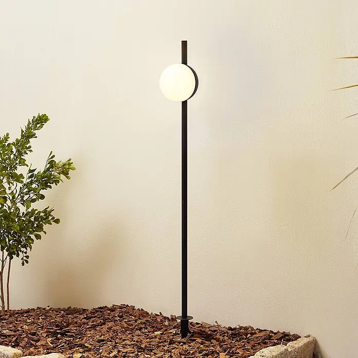 Lindby Erdspießleuchte Darleen -  - Hohe schwarze Bodenspießleuchte mit kugelförmigem weißem Diffusor, in einem Gartenbeet installiert.