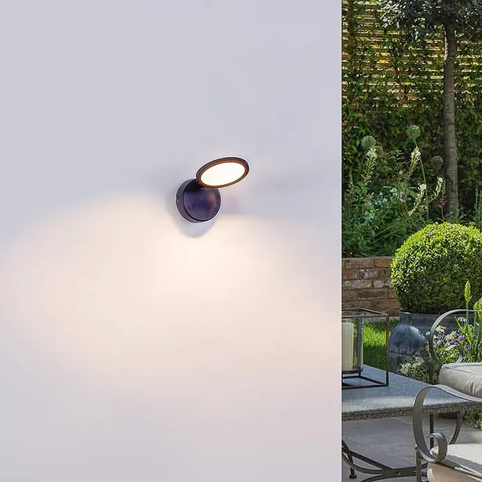 Lindby Deckenstrahler Manel - schwarz, schwarz - Anthrazitfarbene, verstellbare LED-Wandleuchte wirft warmes Licht an eine Außenwand neben einem Garten.