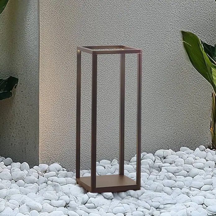 Lucande Außensockelleuchte Thorvid -  - Dunkelbraune rechteckige Outdoor-Laterne oder Deko-Ständer aus Metall auf weißen Kieselsteinen vor einer Wand und Pflanze.