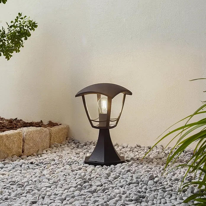 Lindby Außensockelleuchte Isidoro - 16,4 x 16,4 x 28 cm - Dunkle Außenpollerleuchte mit klaren Scheiben und warmer Glühbirne, beleuchtet einen Kiesweg im Garten.