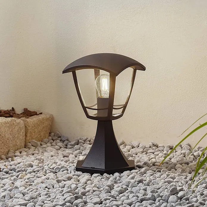 Lindby Außensockelleuchte Isidoro - 16,4 x 16,4 x 28 cm - Dunkle Metall-Gartenlampe mit Klarglas und freiliegender Glühbirne, auf grauen Kieselsteinen.