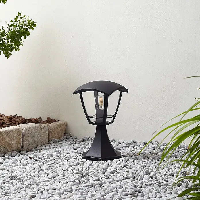 Lindby Außensockelleuchte Isidoro - 16,4 x 16,4 x 28 cm - Schwarze Gartenlampe mit klaren Glasscheiben und sichtbarer Glühbirne auf hellgrauen Kieselsteinen.