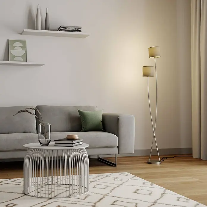 Lucande Stehleuchte Juljana - alu / grau / zink, creme / amber, E14 - Moderne Stehlampe mit zwei hellbeigen Stoffschirmen und sich kreuzenden silbernen Metallstangen, die ein Wohnzimmer beleuchtet.