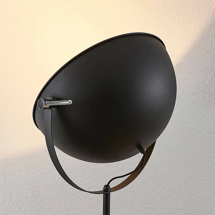 Lindby Stehleuchte Muriel - Höhe: 165 cm, schwarz, schwarz, E27 - Nahaufnahme eines mattschwarzen Metall-Lampenkopfes auf einem Ständer, der warmes Licht auf eine strukturierte Wand wirft.