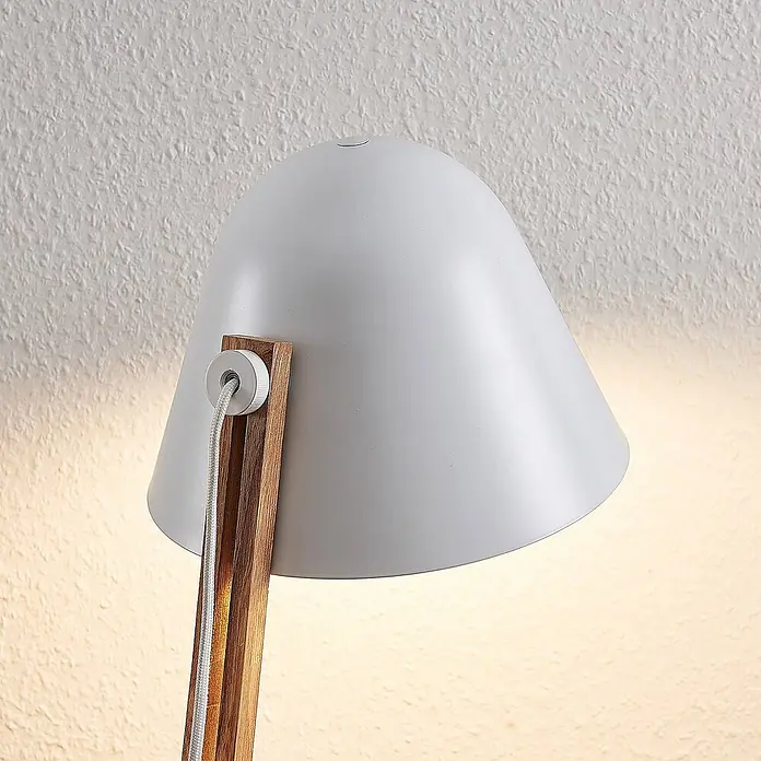 Lindby Stehleuchte Tetja - Höhe: 128 cm, Holz hell, weiß / opal, E27 - Grauer Metall-Lampenschirm mit Holzarm und geflochtenem Kabel, der warmes Licht auf eine strukturierte Wand wirft.