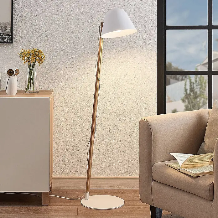 Lindby Stehleuchte Tetja - Höhe: 128 cm, Holz hell, weiß / opal, E27 - Stehleuchte aus weißem Metall und hellem Holz mit konischem Schirm, die einen gemütlichen Wohnbereich neben einem Sofa beleuchtet.