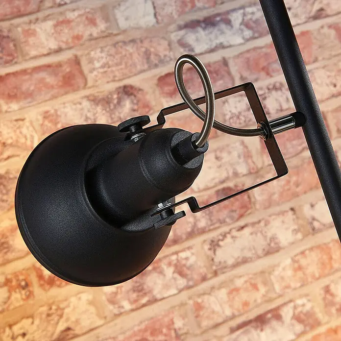 Lindby Stehleuchte Lilly - Höhe: 170 cm, schwarz, schwarz, E14 - Nahaufnahme eines schwarzen Metallstrahlers mit verstellbarem Kopf und flexiblem Metallarm, an einem Ständer vor einer Ziegelwand montiert.
