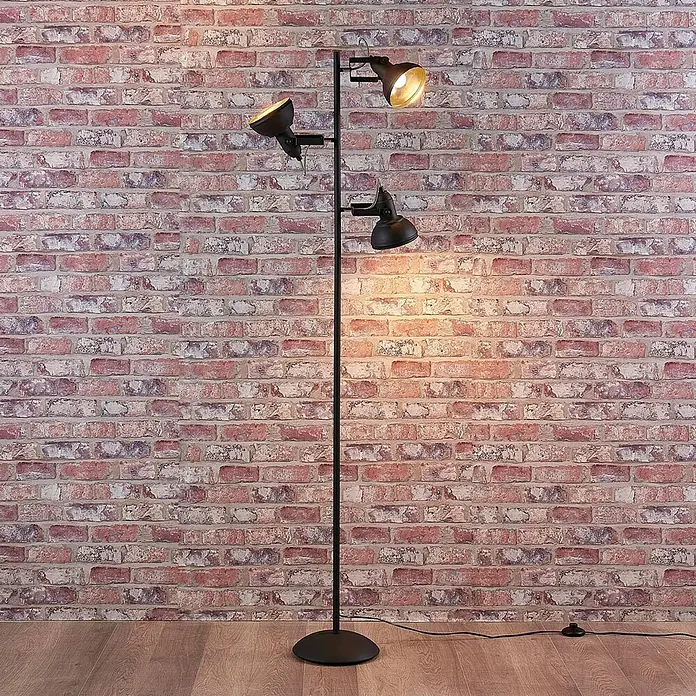 Lindby Stehleuchte Lilly - Höhe: 170 cm, schwarz, schwarz, E14 - Eine schwarze Stehlampe aus Metall mit drei verstellbaren Strahlern, beleuchtet eine Ziegelwand und einen Holzfußboden.
