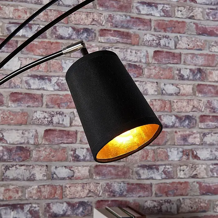 Lindby Stehleuchte Tinne - Höhe: 210 cm, schwarz, schwarz, E14 - Nahaufnahme eines schwarzen Stofflampenschirms mit goldener Innenseite, beleuchtet, an einem schwarzen Metallarm vor einer Ziegelwand.