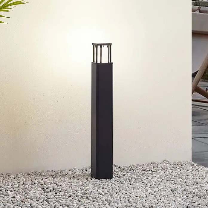 Lucande Außenlaterne Dakil -  - Schwarze, rechteckige Außenpollerleuchte mit offenem Käfigaufsatz, auf Kieselsteinen an einer Wand.