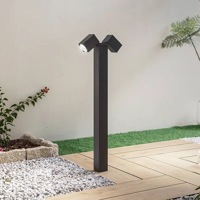 Lindby Außenlaterne Othil - 6,5 x 6,5 x 80 cm - Dunkelgraue Außenpollerleuchte mit zwei verstellbaren Würfelstrahlern, die einen Gartenweg mit Holzdeck und Kieselsteinen beleuchtet.