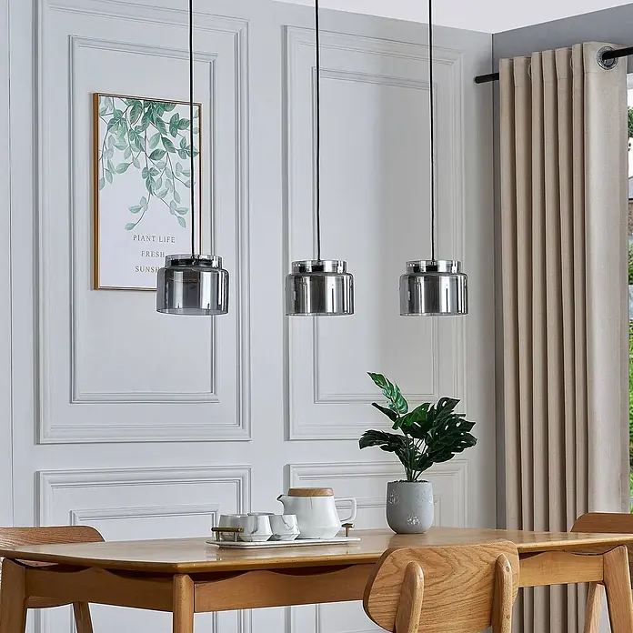 Lucande Pendelleuchte Diano - alu / grau / zink, rauchgrau, chrom - Drei moderne Pendelleuchten mit Rauchglas-Schirmen und Metalloberteilen hängen über einem Holzesstisch.