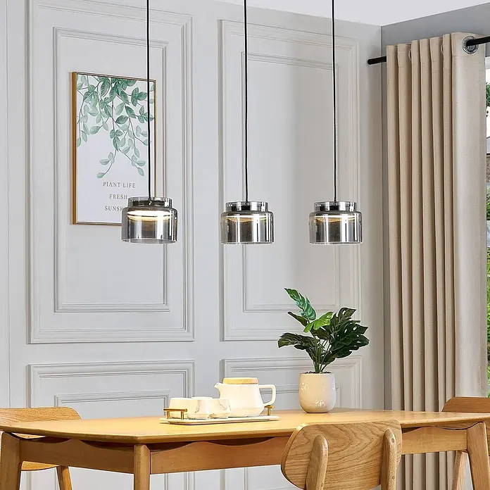 Lucande Pendelleuchte Diano - alu / grau / zink, rauchgrau, chrom - Drei moderne Pendelleuchten mit Rauchglas und Chromakzenten hängen über einem Holzesstisch.