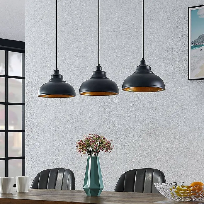 Lindby Pendelleuchte Emna - L x B x H: 101 x 29 x 16 cm, schwarz, Schwarz, gold, E27 - Drei schwarze Industrie-Pendelleuchten mit goldenem Innenleben hängen über einem Holzesstisch.