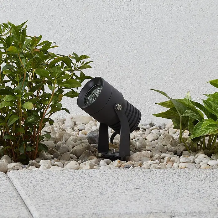 Lucande LED Gartenstrahler mit Erdspieß in dunkelgrau, Strahler Garten -  - Schwarzer Outdoor-Strahler zwischen Kieselsteinen und Pflanzen, beleuchtet einen Gartenweg.