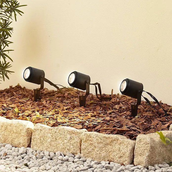 Lindby Erdspießleuchte Nymo -  - Drei schwarze Gartenstrahler in einem Beet mit Rindenmulch, die eine Wand beleuchten.