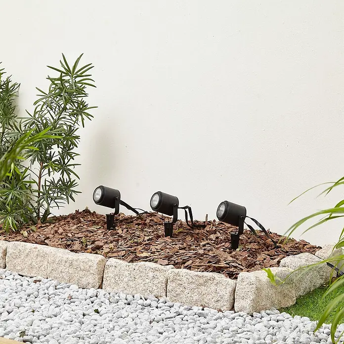 Lindby Erdspießleuchte Nymo -  - Drei schwarze Gartenstrahler in Rindenmulch, neben einem Steinrand und weißen Kieselsteinen.