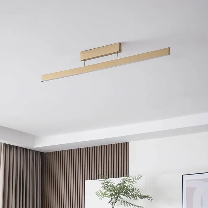 Lucande Deckenleuchte Mylosh - messing / gold - Eine goldfarbene lineare LED-Deckenleuchte in einem modernen Wohnraum installiert, die den Raum beleuchtet.