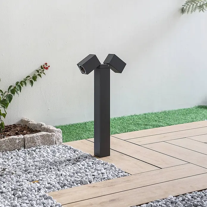 Lindby Außensockelleuchte Othil -  - Schwarze Außenleuchte mit zwei verstellbaren Strahlern, platziert in einem Garten mit Kies und Holzterrasse.