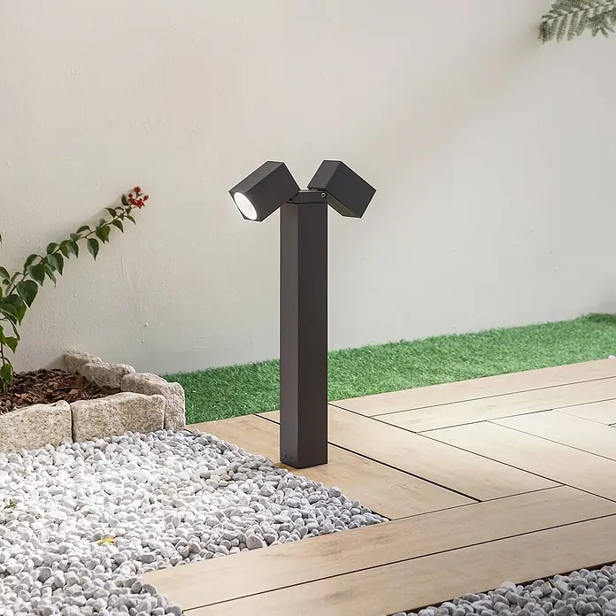 Lindby Außensockelleuchte Othil -  - Dunkelgraue Außenpollerleuchte mit zwei verstellbaren Köpfen, die einen Gartenweg mit Kies und Holzterrassen beleuchtet.