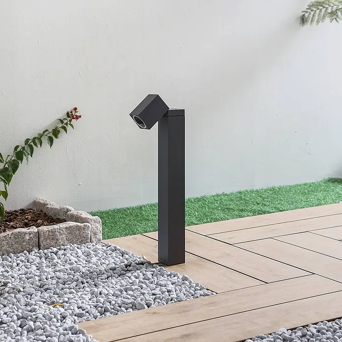 Lindby Außensockelleuchte Othil -  - Dunkelgraue Außenpollerleuchte mit verstellbarem Kopf, die einen Gartenweg mit Kies und Holzstegen beleuchtet.