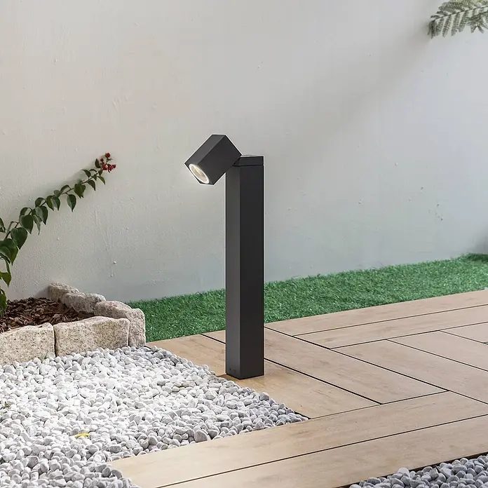 Lindby Außensockelleuchte Othil -  - Dunkelgraue Outdoor-LED-Pollerleuchte mit angewinkeltem Kopf, die eine Gartenmauer beleuchtet.