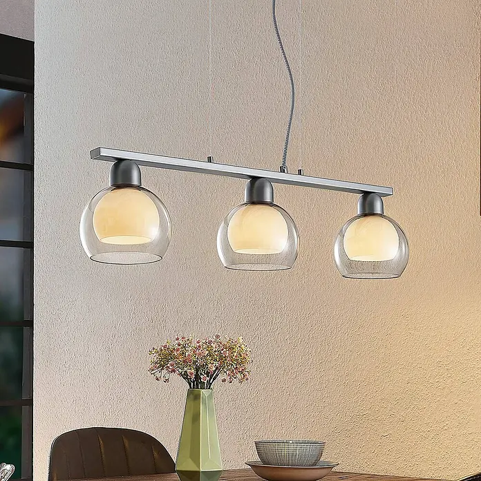 Lucande Pendelleuchte Kaiya - L x B x H: 68 x 14 x 7,8 cm, nickel, weiß, rauchgrau, E14 - Lineare Pendelleuchte mit drei Kugelgläsern aus Klar- und Opalglas an einer Metallstange, die einen Esstisch beleuchtet.
