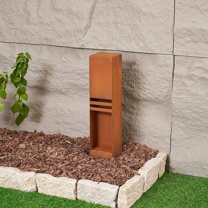 Lucande Außensockelleuchte Oxido - 12 x 12 x 50 cm - Rostfarbene Gartenleuchte in einem Gartenbeet mit Rindenmulch und Steinumrandung.