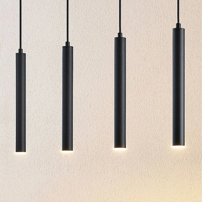 Arcchio Pendelleuchte Kammeron - schwarz, Schwarz - Vier schwarze zylindrische Pendelleuchten hängen vor einer strukturierten Wand und beleuchten diese nach unten mit warmem Licht.