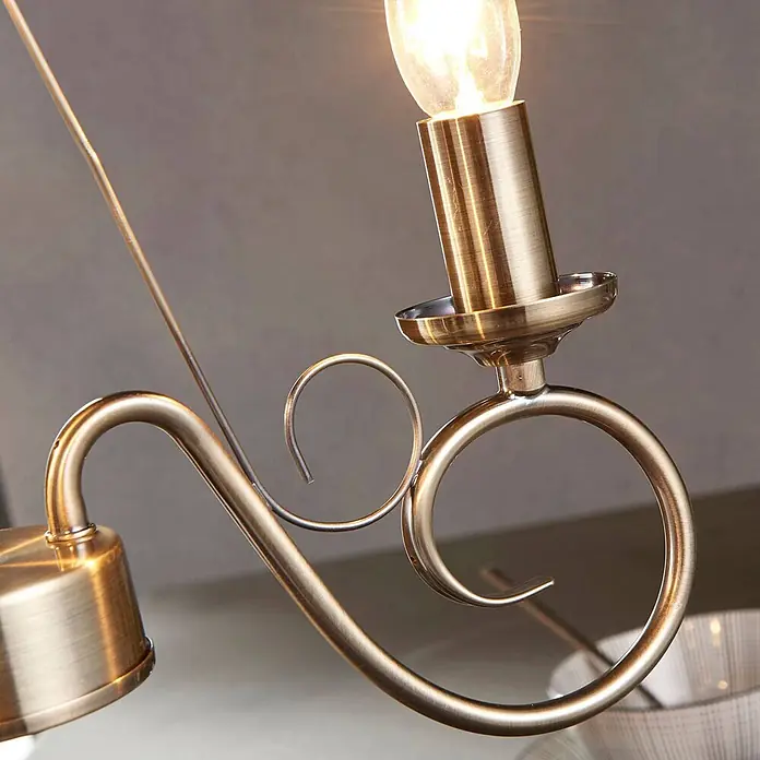 Lindby Kronleuchter Marnia - bronze / altmessing, altmessing, E14 - Detailaufnahme einer Leuchte aus Antikmessing mit leuchtender Birne und dekorativen Metallornamenten.