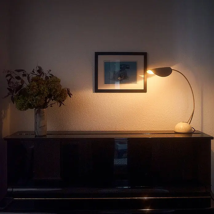 Lucande Tischleuchte Rivera - alu / grau / zink, alu / grau / zink, G9 - Eine cremefarbene, gebogene Tischlampe mit dunklem, blattförmigem Schirm beleuchtet ein gerahmtes Bild und die Wand über einem dunklen Klavier.