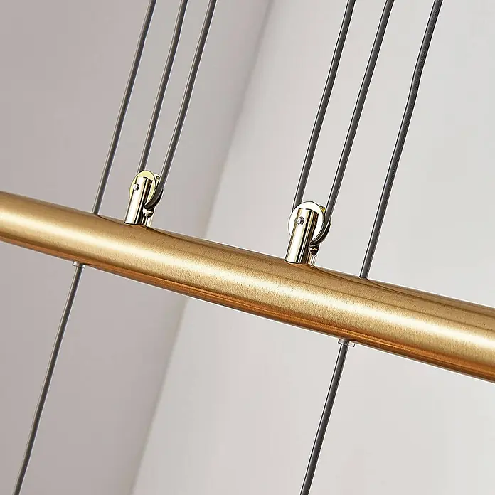 Lindby Pendelleuchte Simeon - L x B x H: 72,5 x 13 x 12 cm, weiß, messing matt, E14 - Nahaufnahme einer gebürsteten Goldstange mit dunklen Kabeln, die von kleinen Goldklammern gehalten werden.