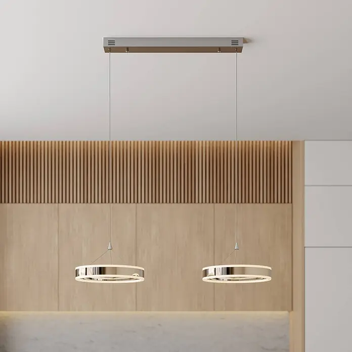Lucande Pendelleuchte Lyani - L x B x H: 85 x 30 x 5 cm, chrom / silber, chrom, weiß satiniert - Zwei moderne Chrom-Pendelleuchten mit beleuchteten Ringen hängen von einer rechteckigen Deckenhalterung über einer Holzwand.