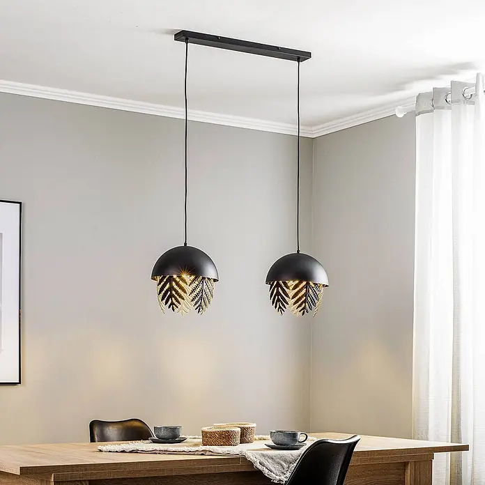 Lucande Pendelleuchte Aparas - L x B x H: 84 x 28 x 35 cm, schwarz, Schwarz, gold, E27 - Zwei schwarze Pendelleuchten mit goldenen blattgemusterten Schirmen über einem Esstisch.