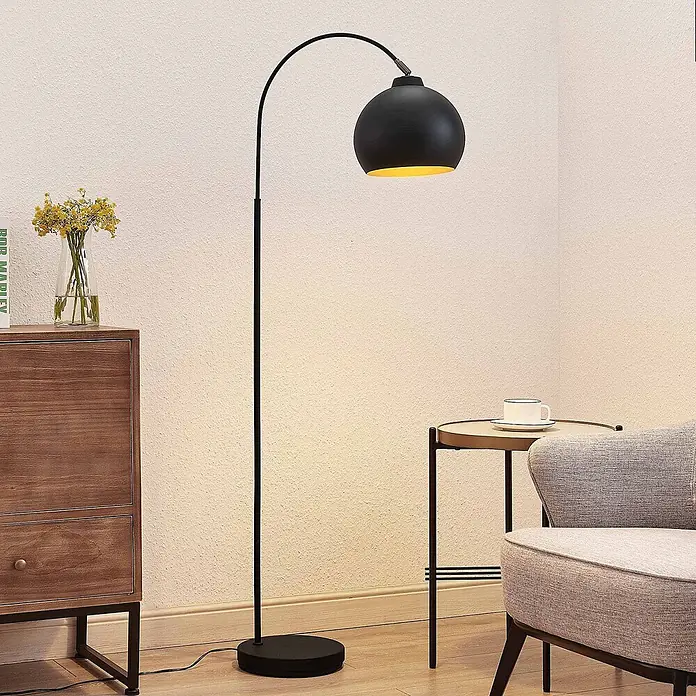 Lindby Stehleuchte Minali - Höhe: 148 cm, schwarz, schwarz, E27 - Schwarze Bogen-Stehleuchte mit kugelförmigem Schirm und goldener Innenseite, die warmes Licht neben einem Sessel spendet.