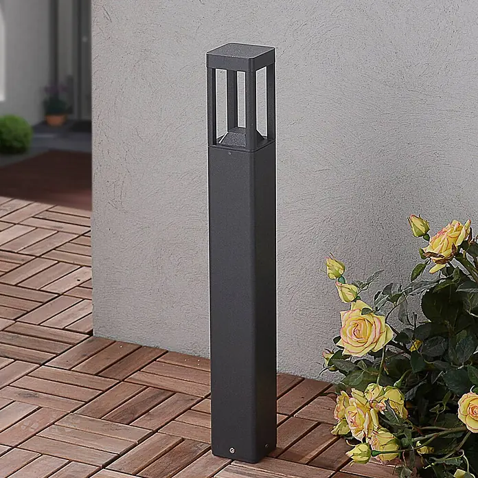 Lucande Außenlaterne Bernd -  - Dunkelgraue Pollerleuchte mit offenem Design, auf Holzfliesen neben einer Wand und gelben Rosen.