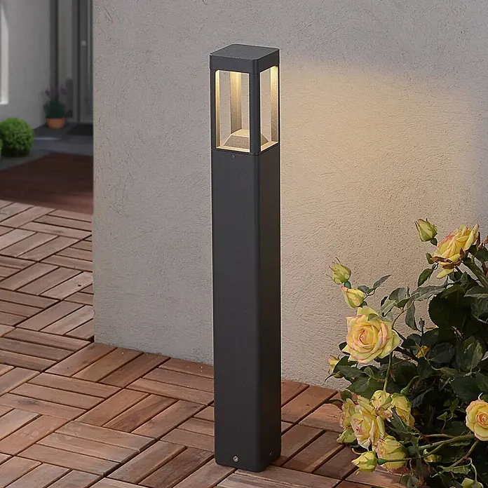 Lucande Außenlaterne Bernd -  - Dunkelgraue, quadratische Außenpollerleuchte auf Holzterrassendielen, strahlt warmes Licht aus.