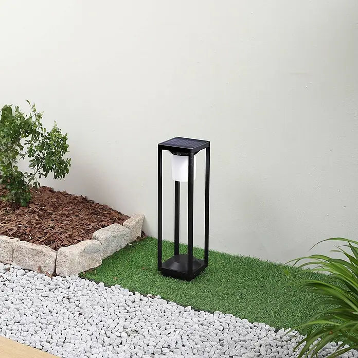 Lucande Außenlaterne Nilea -  - Schwarze Solar-Gartenleuchte mit weißem Diffusor, auf Kunstrasen in einem Garten neben einem weißen Kiesweg stehend.