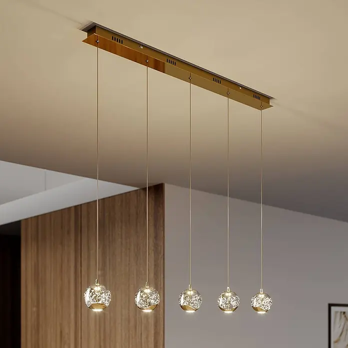 Lucande Pendelleuchte Hayley - L x B x H: 96 x 9 x 9 cm, klar / transparent, klar, gold - Fünf gold-akzentuierte, kugelförmige Pendelleuchten mit funkelndem Inneren hängen an einer rechteckigen goldenen Deckenleiste.