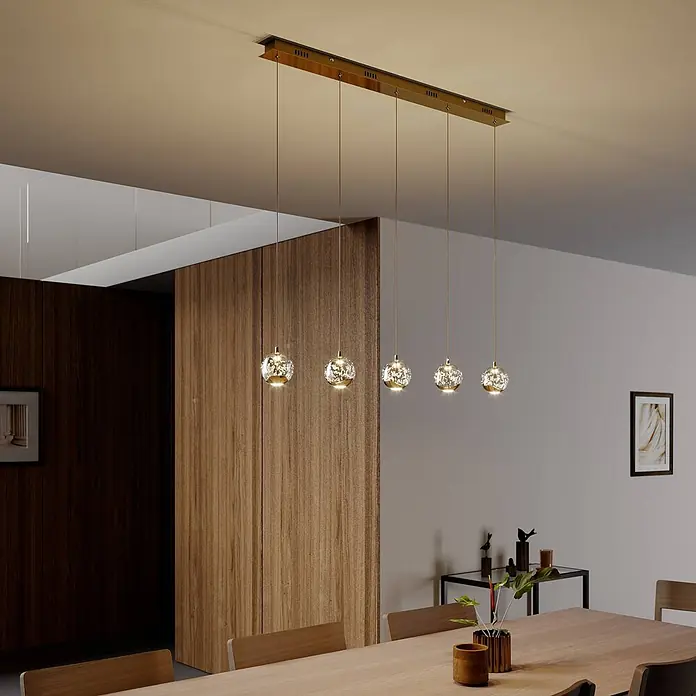 Lucande Pendelleuchte Hayley - L x B x H: 96 x 9 x 9 cm, klar / transparent, klar, gold - Goldene lineare Pendelleuchte mit fünf funkelnden Kristallkugeln über einem Holztisch.