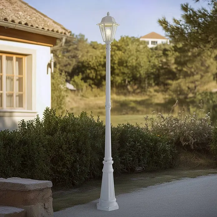 Lindby Außenlaterne Edana - 23 x 26 x 211 cm - Weißer Gartenlaternenpfahl mit klassischem Laternenkopf und leuchtender Glühbirne, an einem Gartenweg neben einem Haus.