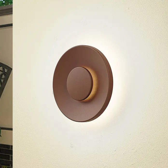 Lucande Außenwandleuchte Kayana - braun / rost, IP65 - Eine runde, dunkelbronzene Wandleuchte mit erhöhter Mitte, die ein warmes Licht an eine strukturierte Wand abgibt.