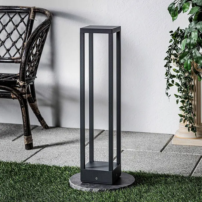 Lucande Außenlaterne Carlota -  - Hohe, schwarze, rechteckige Außenleuchte mit offenem Rahmendesign auf einem Steinsockel auf einer Terrasse.