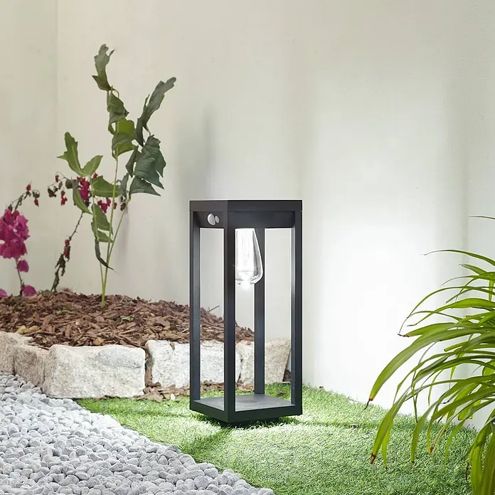 Lindby Außenlaterne Enea -  - Schwarze, rechteckige Außenleuchte mit sichtbarer Birne, beleuchtet einen Gartenweg auf Kunstrasen.