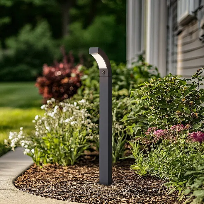 Lucande Außenlaterne Lennik - 20 x 12 x 100 cm - Dunkelgraue moderne Wegeleuchte mit gebogenem Kopf und integriertem Sensor, beleuchtet ein Gartenbeet.