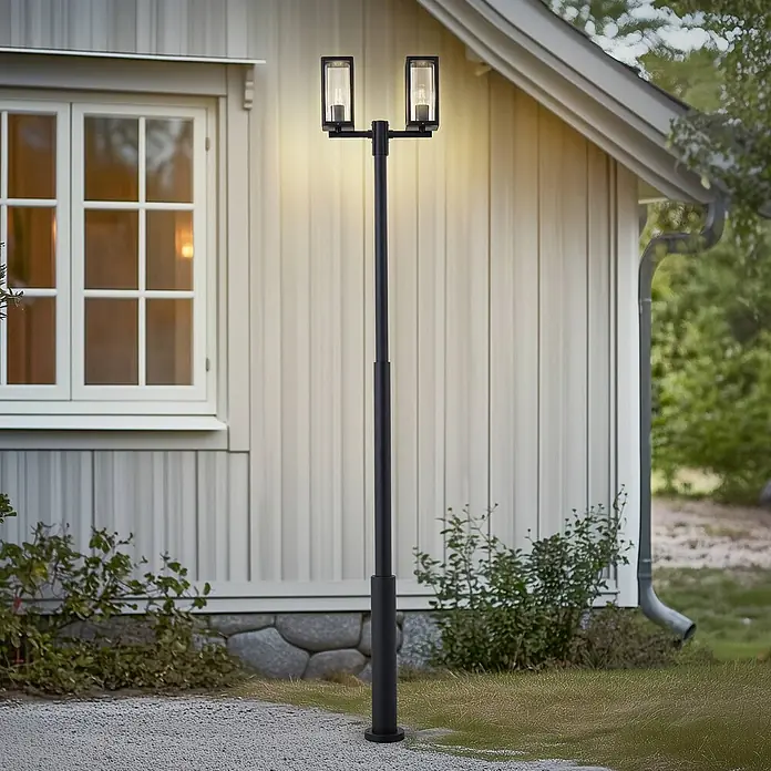 Lucande Außenlaterne Siveta -  - Schwarze Metall-Gartenleuchte mit zwei rechteckigen Lampen beleuchtet einen Garten neben einem Haus.