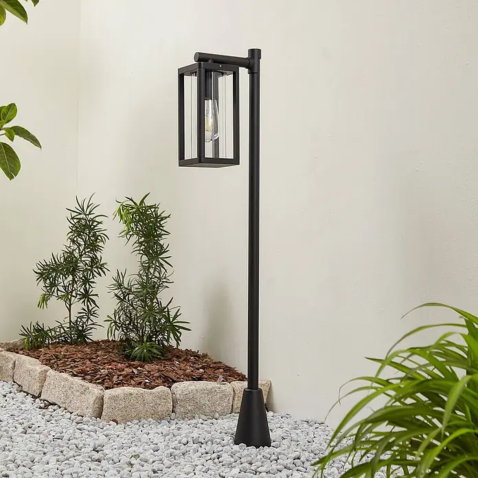 Lucande Außenlaterne Siveta -  - Moderne schwarze Außenstandleuchte mit klarem Glaslaternenkopf und freiliegender Glühbirne im Garten auf weißen Kieselsteinen.