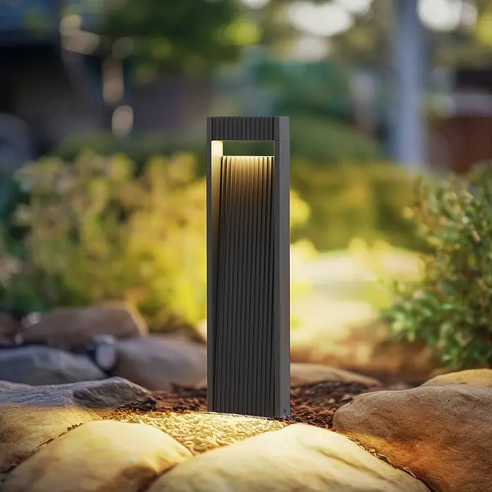 Lucande Außenlaterne Tavion -  - Dunkelgraue, gerippte Gartenwegleuchte, die Felsen mit warmem Licht in natürlicher Umgebung beleuchtet.