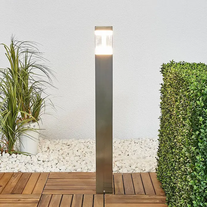 Lindby LED Wegeleuchte in edelstahl, transparent, Gartenlampe -  - Hohe, rechteckige Pollerleuchte aus Edelstahl mit beleuchteter Oberseite, im Außenbereich auf Holzterrasse zwischen weißen Kieselsteinen.